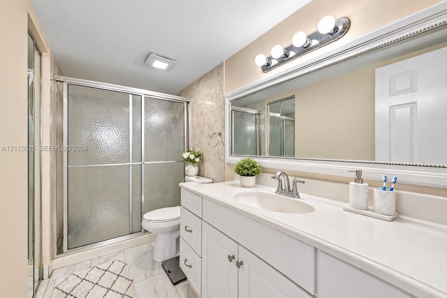 Bathroom featuring a white vanity with a solid-surface countertop, an integrated sink basin, a full-width framed mirror, a glass-enclosed shower with a sliding door, and neutral tile flooring