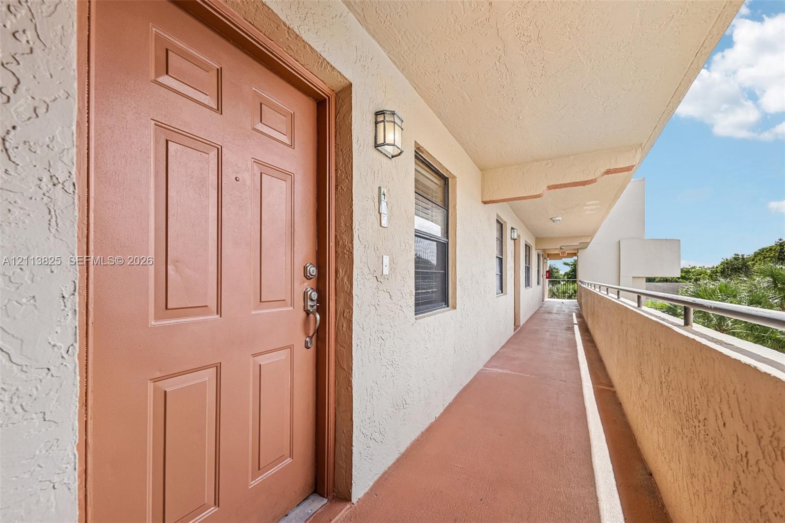 Exterior entry featuring a raised-panel door, textured stucco wall finishes, metal safety railing, and wall-mounted exterior lighting