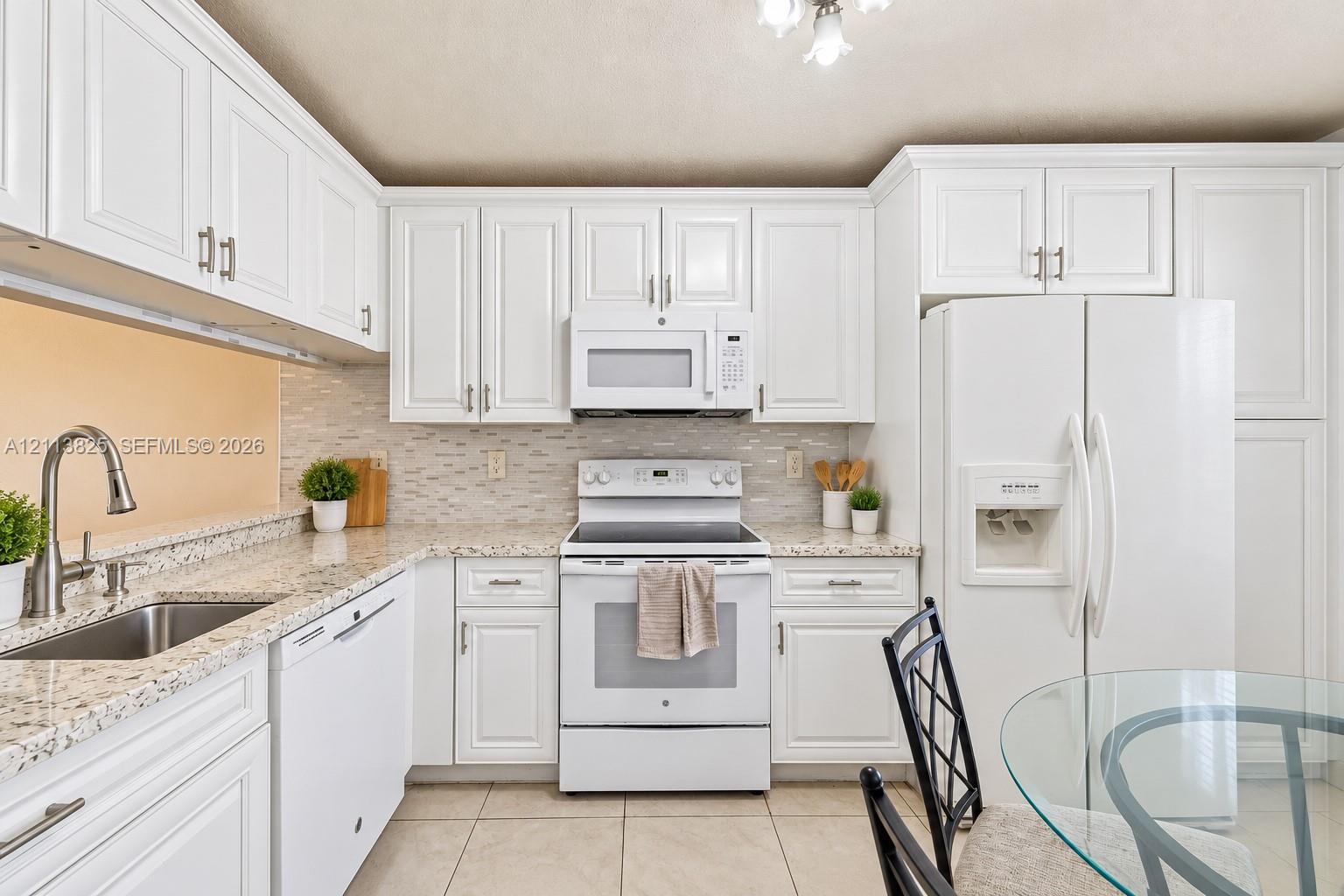 Kitchen featuring white shaker-style cabinetry, speckled stone-finish countertops, a subway-patterned tile backsplash, stainless steel under-mount sink with a high-arch faucet, and white appliances