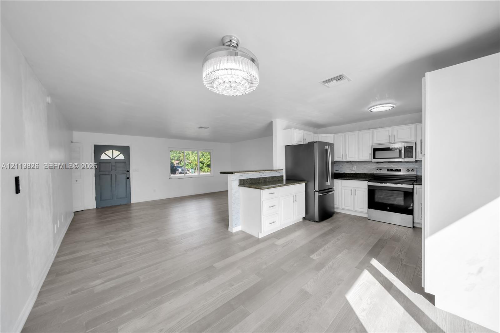 Open-concept kitchen featuring white shaker-style cabinetry, dark stone-finish countertops, stainless steel appliances, and a peninsula island with decorative tile detailing, complemented by wood-finish flooring and a central crystal-style chandelier