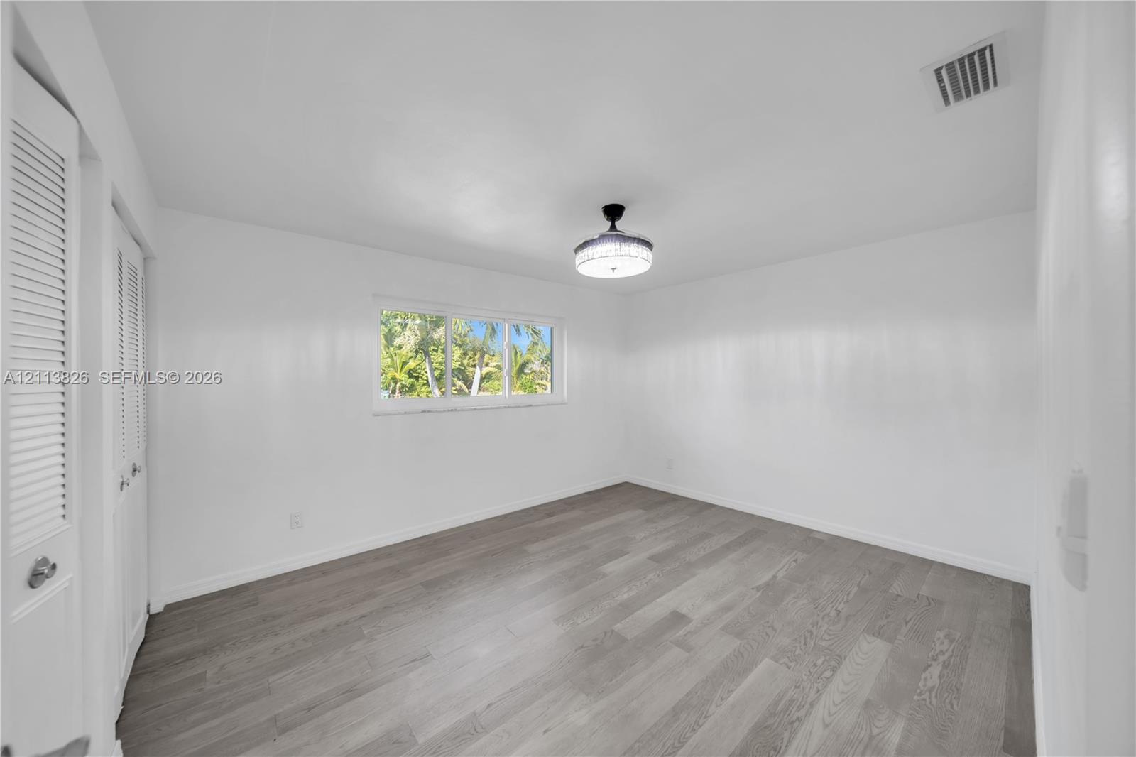 Room featuring wood-finish flooring, white walls, dual louvered-panel closet doors, a central flush-mount light fixture, and a window with exterior greenery