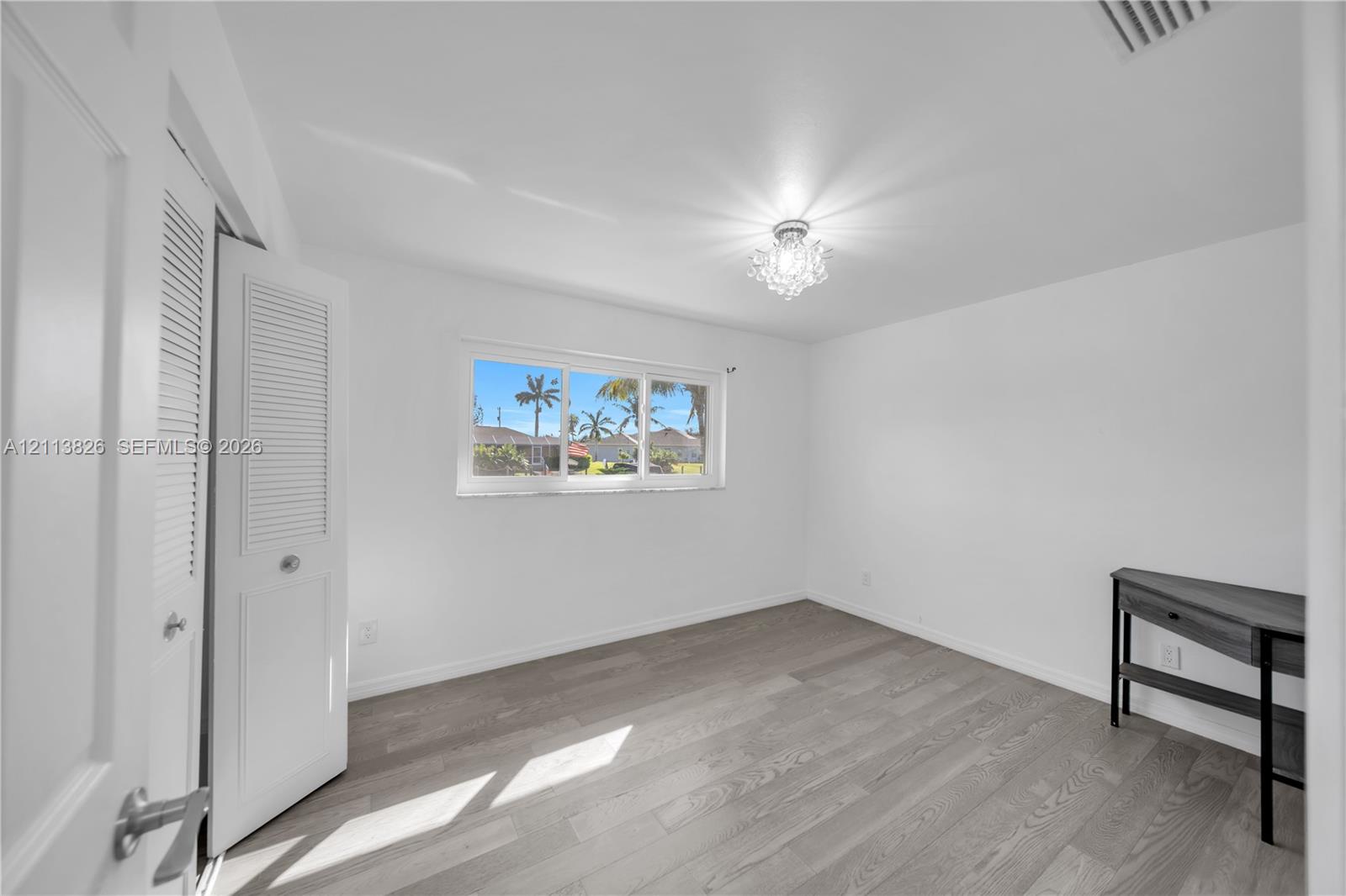 Wood-finish flooring, white louvered closet doors, multi-pane horizontal window, and decorative crystal-style ceiling fixture