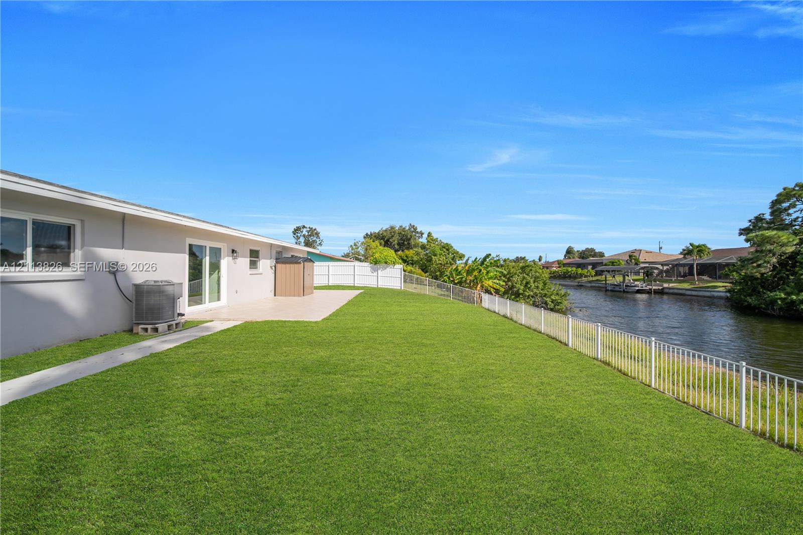 Backyard featuring a concrete patio, perimeter white fencing, manicured lawn, and water frontage