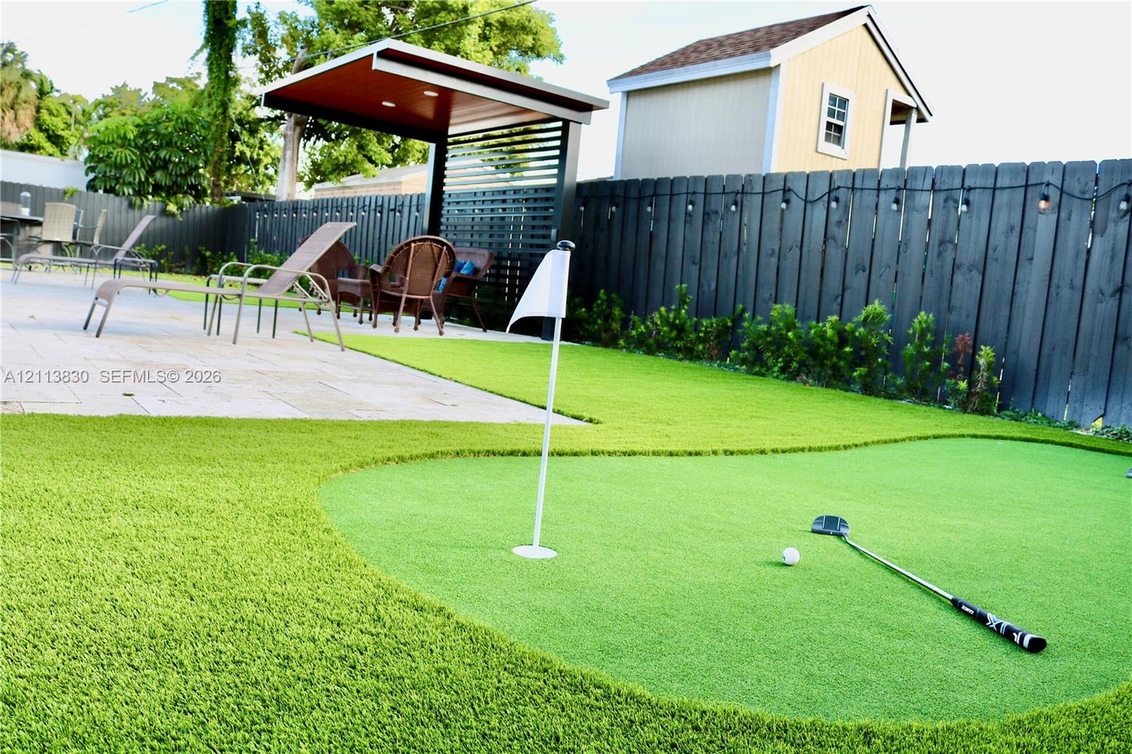 Outdoor putting green featuring synthetic turf, paver patio, covered pavilion with slatted privacy screen, and fenced perimeter