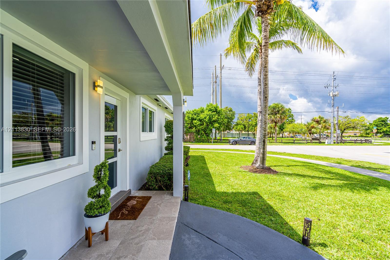 Covered entryway featuring stone-finish floor tiling, exterior wall-mounted lighting, and a glass-paneled entry door, complemented by a manicured lawn and mature palm tree