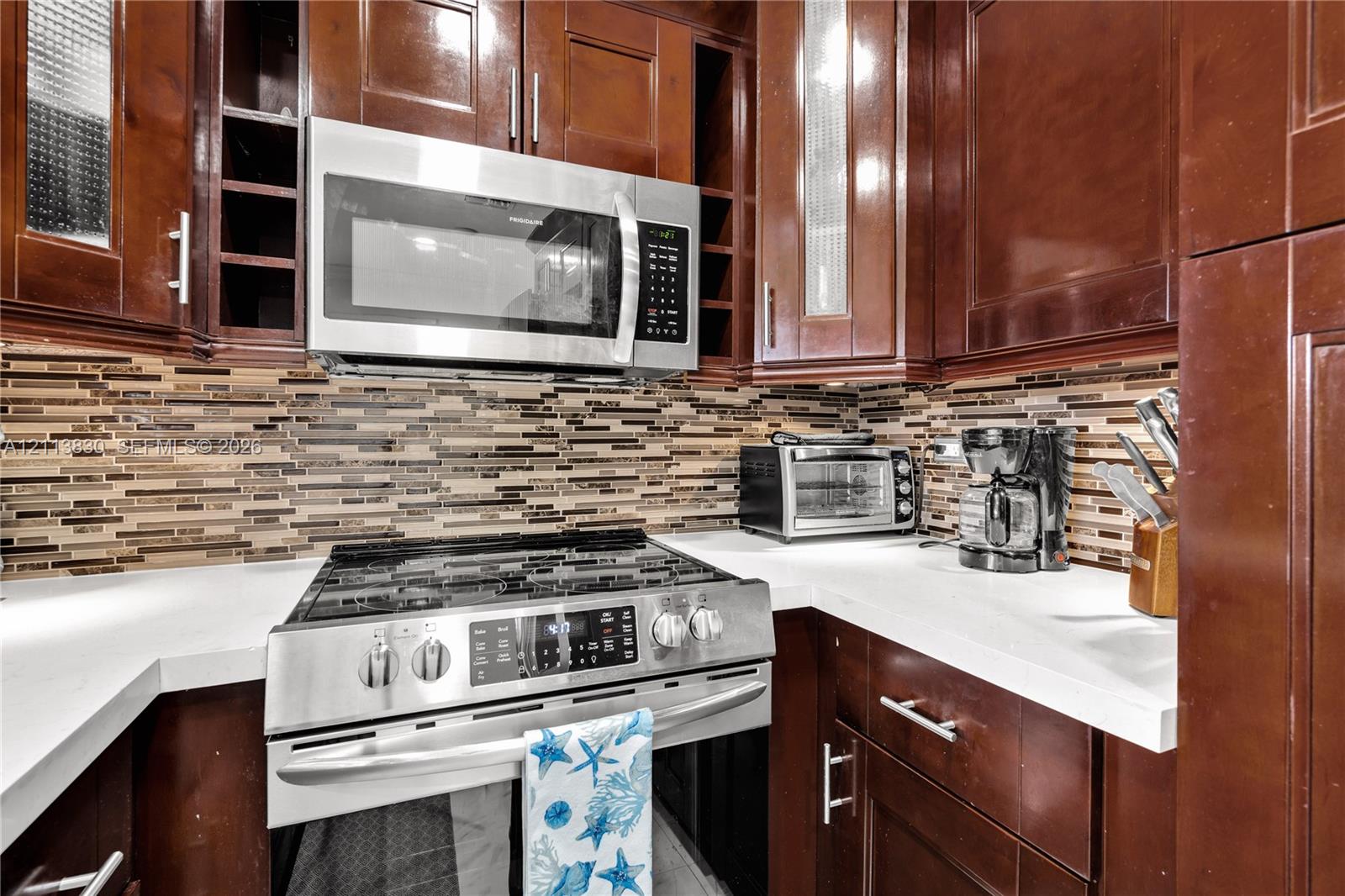 Kitchen featuring dark-finish wood cabinetry, quartz countertops, multi-toned horizontal glass tile backsplash, and stainless steel appliances including a gas range and over-the-range microwave