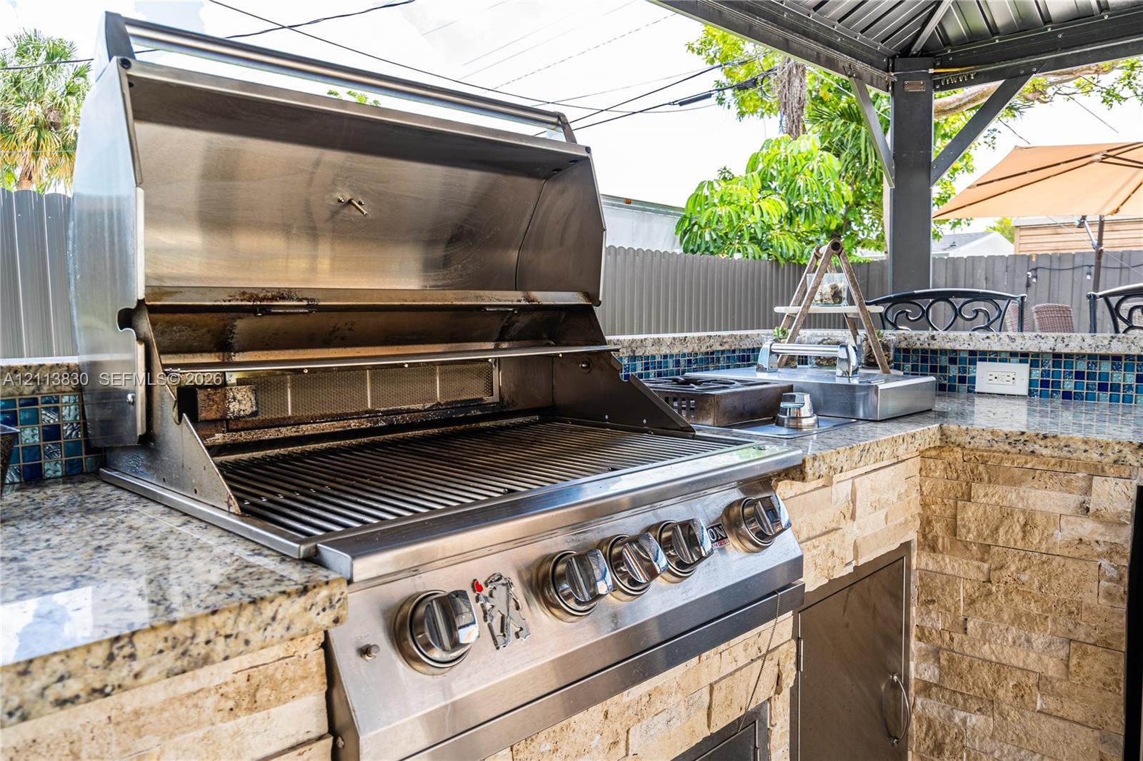 Outdoor kitchen featuring a built-in stainless steel grill, stone veneer base, patterned glass tile backsplash, and stone countertop under a covered structure