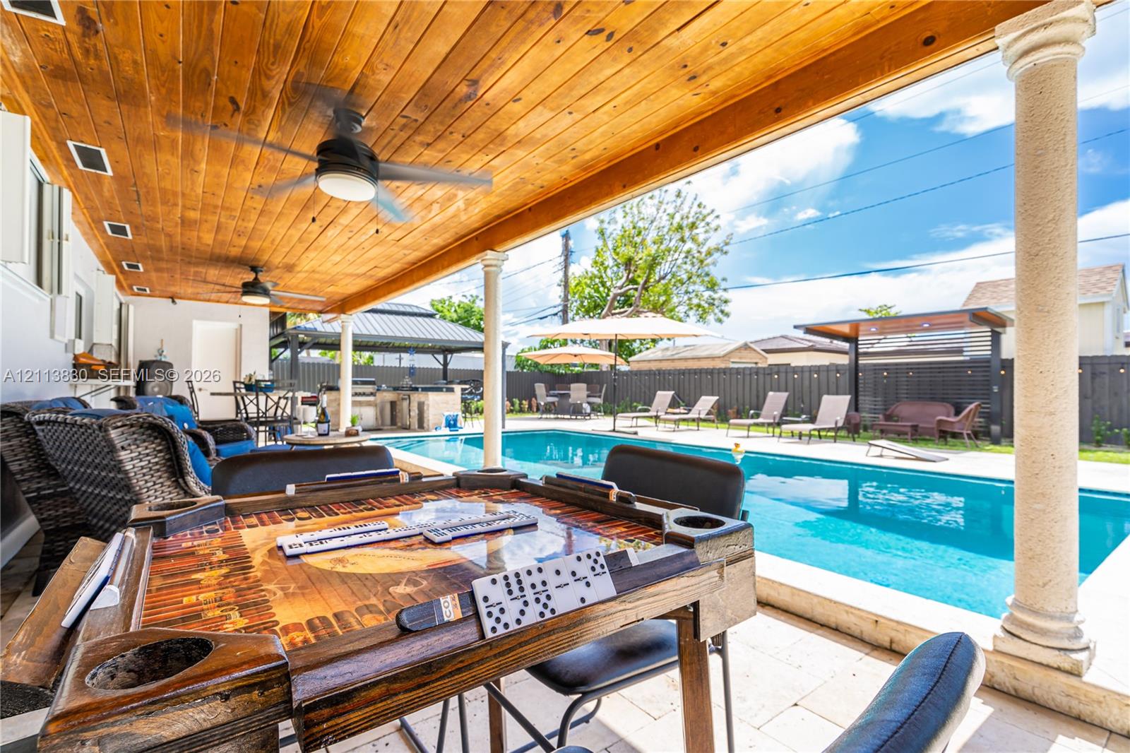 Covered patio featuring a stained wood plank ceiling, recessed lighting, and structural columns overlooking a rectangular swimming pool with paved decking and a built-in outdoor kitchen