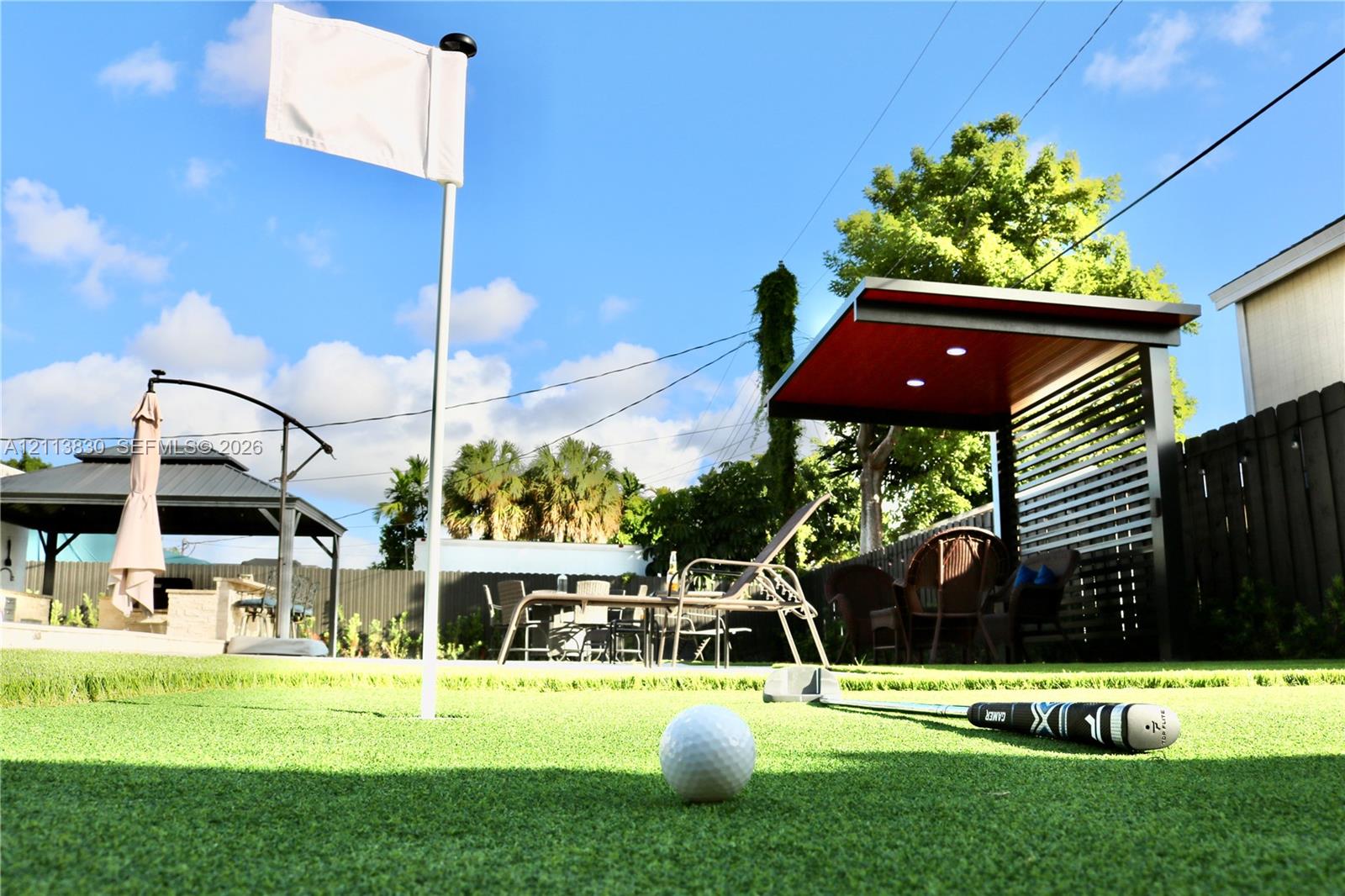 Backyard putting green with integrated flag pole, covered patio structure with slatted privacy screen, and recessed ceiling lighting