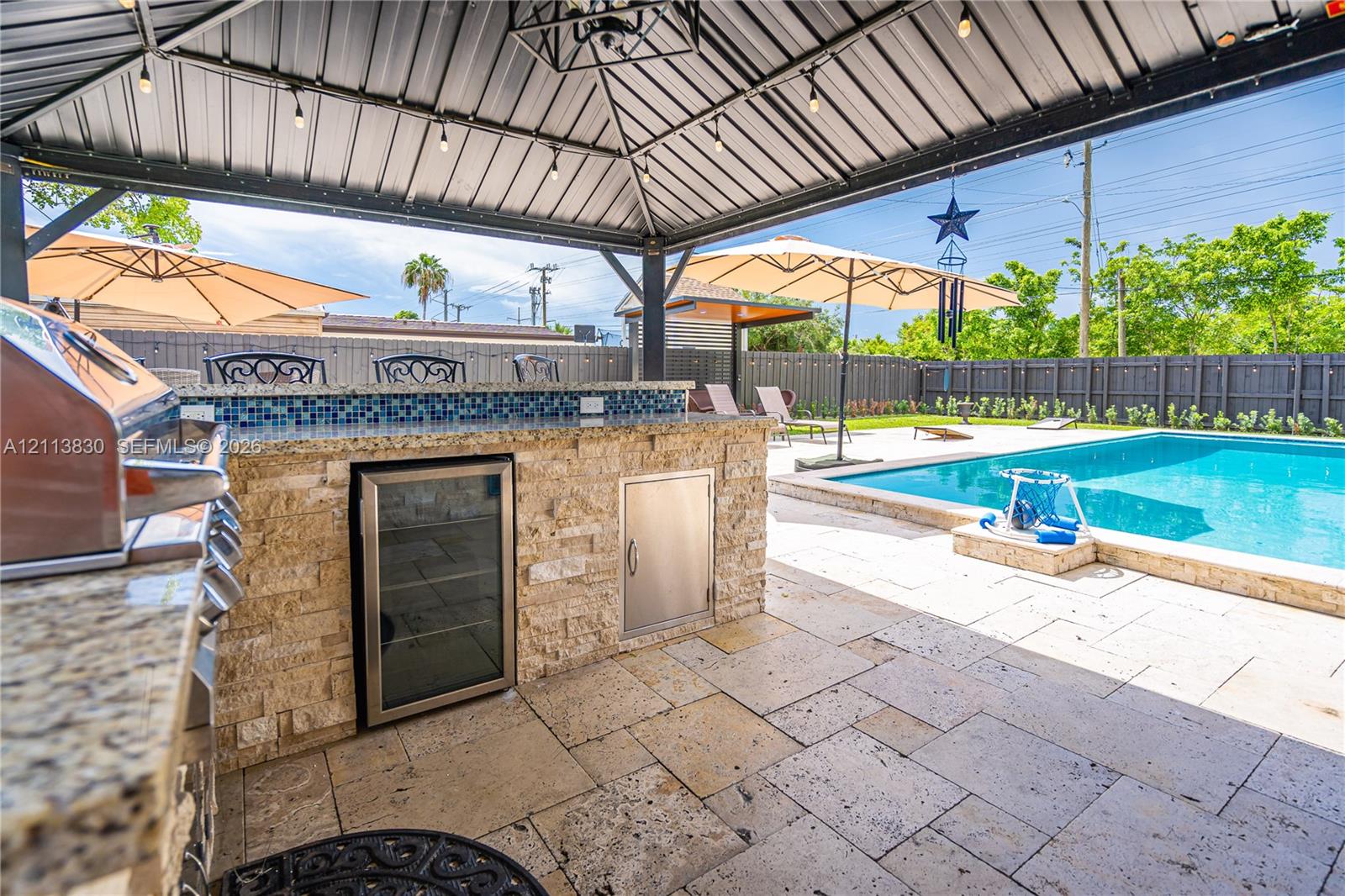 Covered outdoor kitchen featuring stone veneer, integrated stainless steel appliances, and stone-finish countertops, paired with a pool and paved patio