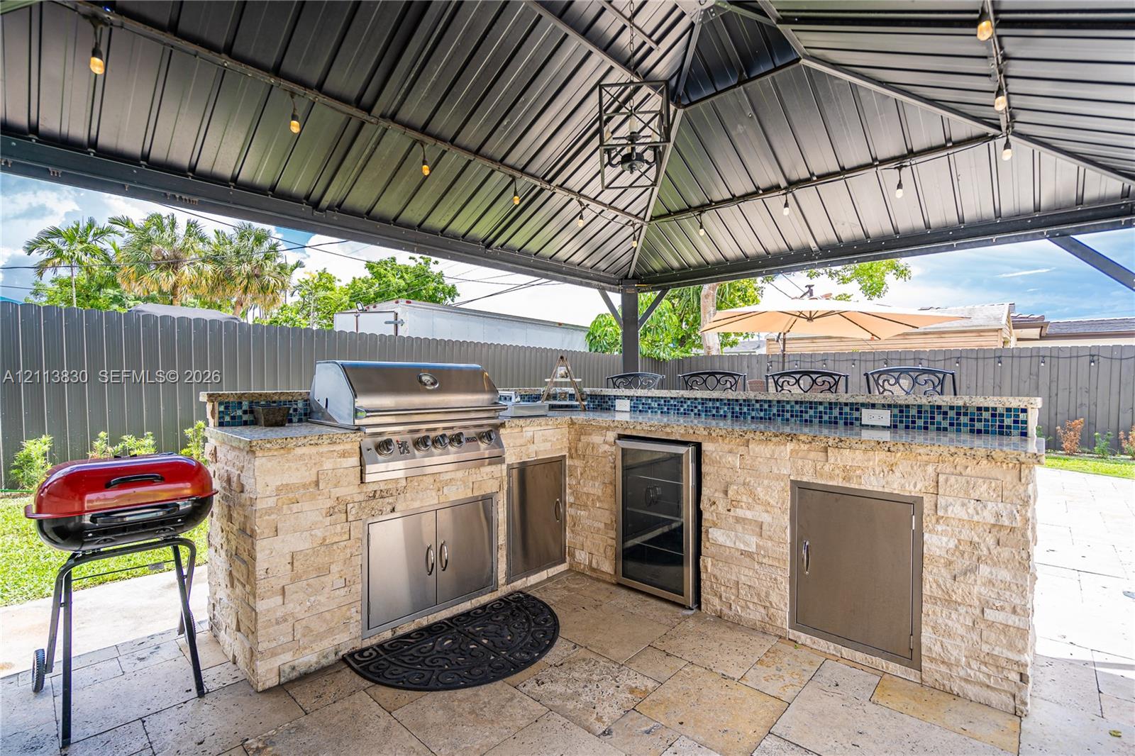Covered outdoor kitchen featuring a stone-clad island, stainless steel grill, beverage refrigerator, tile backsplash, and dark metal gazebo structure