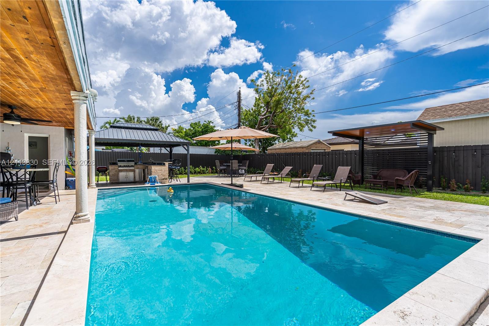 Outdoor area featuring an in-ground swimming pool, stone paver decking, covered patio with wood-paneled ceiling, and a built-in outdoor kitchen with a metal-roof gazebo