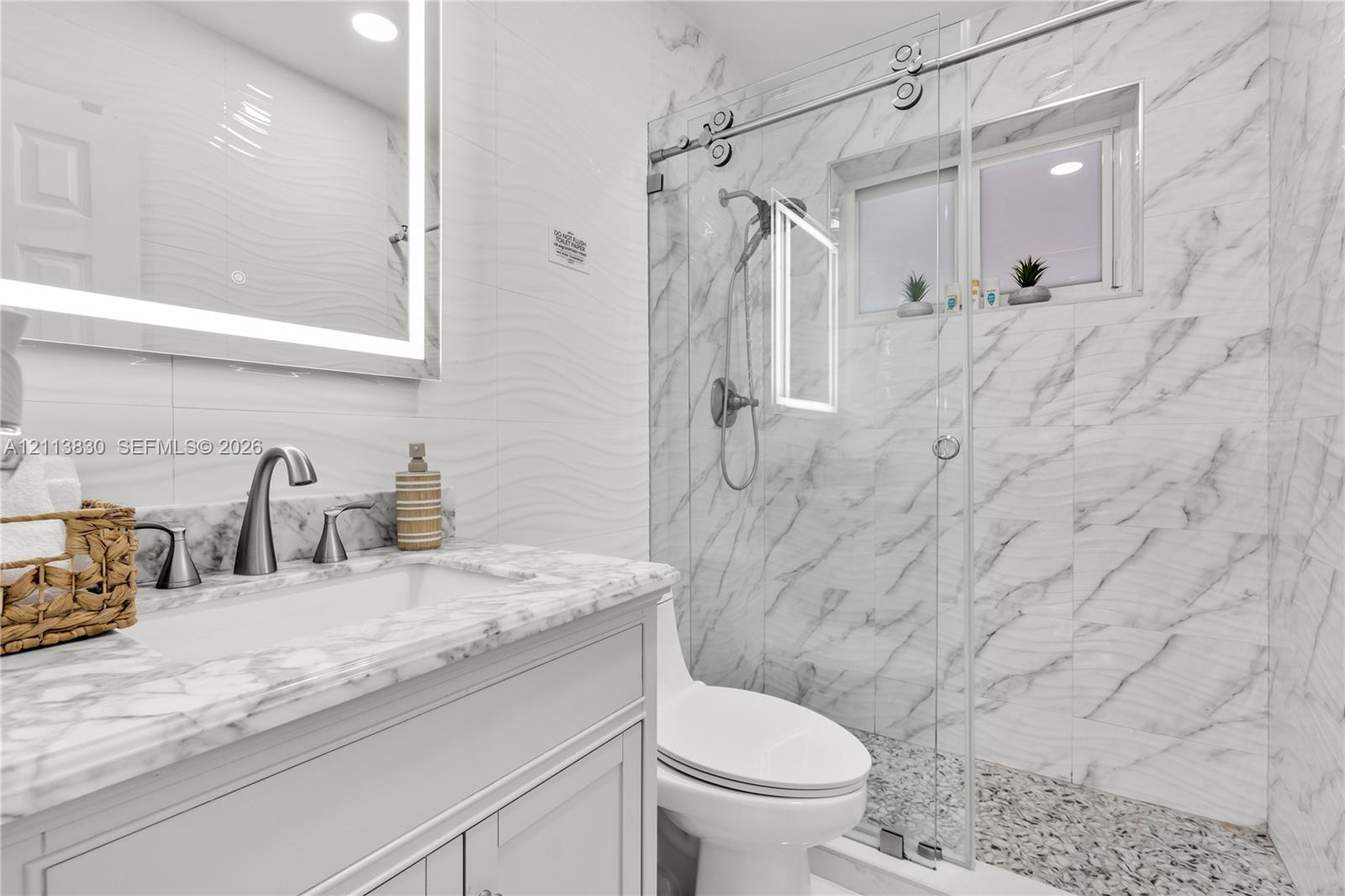 Bathroom featuring a vanity with stone-top basin, integrated LED-backlit mirror, wall-mounted faucet, and glass-enclosed shower with tiled surround and textured flooring