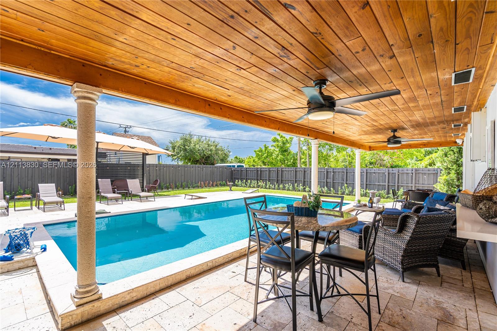 Covered patio featuring a stained wood plank ceiling, dual ceiling fans, stone paver decking, and an in-ground swimming pool with a stone coping border