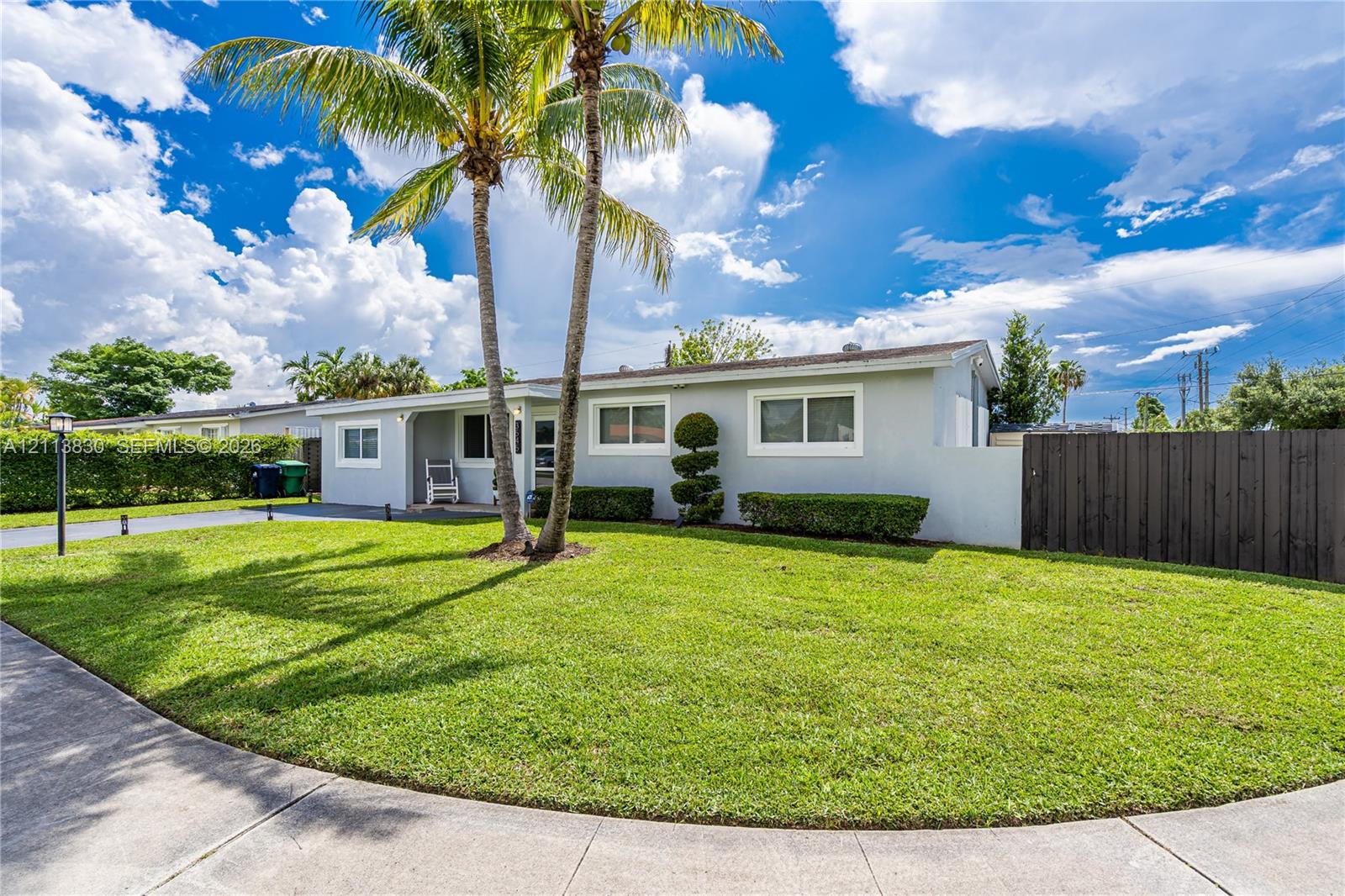 Single-story exterior featuring a light-toned smooth finish, manicured lawn, tall palm trees, and tall wood fencing