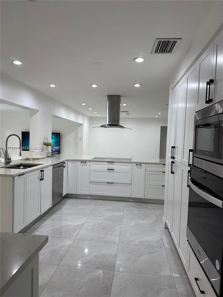 Kitchen featuring white shaker-style cabinetry, stainless steel range hood, built-in double oven, stone-finish countertops, and large-format tile flooring Kitchen featuring white shaker-style cabinetry, stainless steel range hood, built-in double oven, stone-finish countertops, and large-format tile flooring