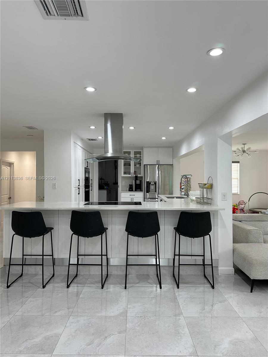 Kitchen featuring white cabinetry, an expansive waterfall-edge peninsula with stone-finish countertops, stainless steel appliances, a chimney-style range hood, and large-format tile flooring Kitchen featuring white cabinetry, an expansive waterfall-edge peninsula with stone-finish countertops, stainless steel appliances, a chimney-style range hood, and large-format tile flooring