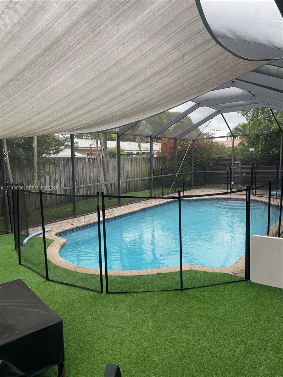 Screened-in pool area featuring a brick paver surround, synthetic turf ground cover, removable black safety fencing, and an overhead sun shade