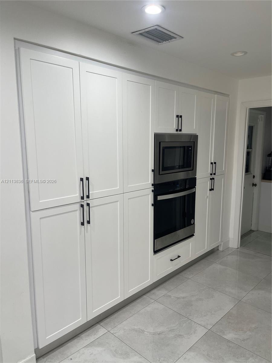 Full-height white cabinetry with Shaker-style doors, matte black hardware, recessed lighting, stainless steel built-in double oven and microwave, and large-format porcelain tile flooring
