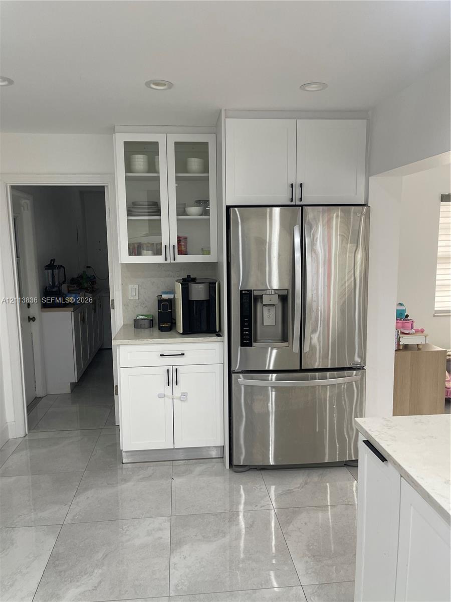 Kitchen featuring white shaker-style cabinetry with glass-front uppers, stainless steel refrigerator, stone-composite countertops, and large-format porcelain tile flooring