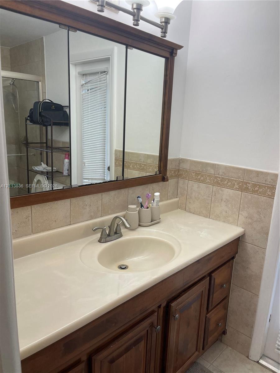 Bathroom vanity featuring wood-finish cabinetry, a molded countertop with integrated basin, polished metal faucet, tri-fold medicine cabinet, and tiled backsplash with decorative inlay