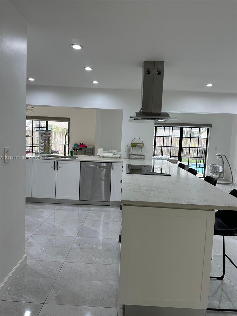 Kitchen featuring a large stone-surface island, white cabinetry, stainless steel dishwasher, stainless steel range hood, and recessed lighting Kitchen featuring a large stone-surface island, white cabinetry, stainless steel dishwasher, stainless steel range hood, and recessed lighting