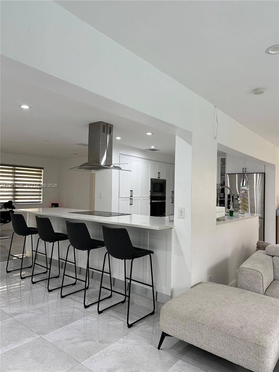 Kitchen featuring white shaker-style cabinetry, an expansive peninsula with a stone-finish countertop, a stainless steel chimney-style range hood, and large-format tile flooring