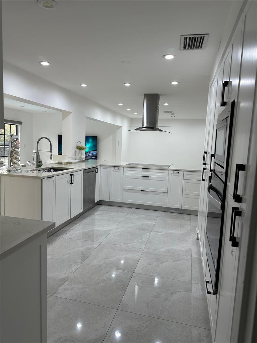 Kitchen featuring white shaker cabinetry, stone-finish countertops, stainless steel appliances including a built-in wall oven and range hood, and large-format porcelain tile flooring Kitchen featuring white shaker cabinetry, stone-finish countertops, stainless steel appliances including a built-in wall oven and range hood, and large-format porcelain tile flooring