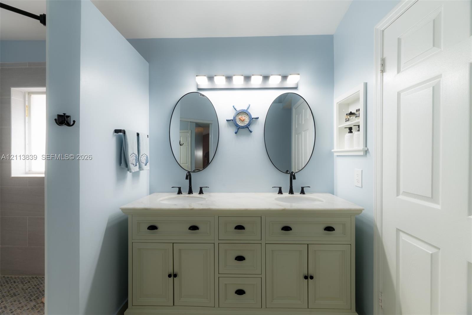 Double vanity featuring stone-finish countertop, dual oval mirrors, recessed wall shelving, and dark-finish hardware