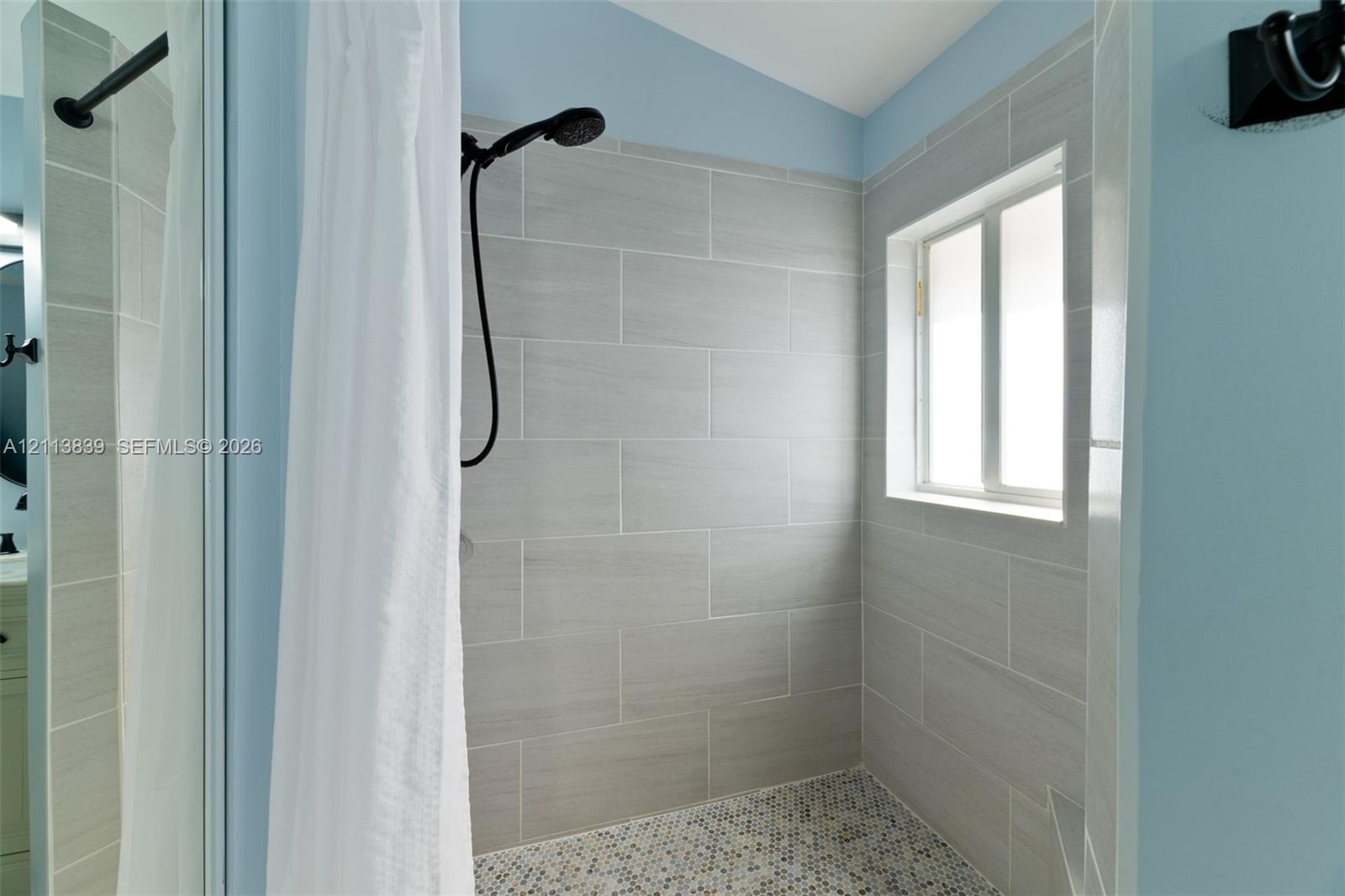 Tiled shower surround with matte black hardware, pebble-stone flooring, and inset window