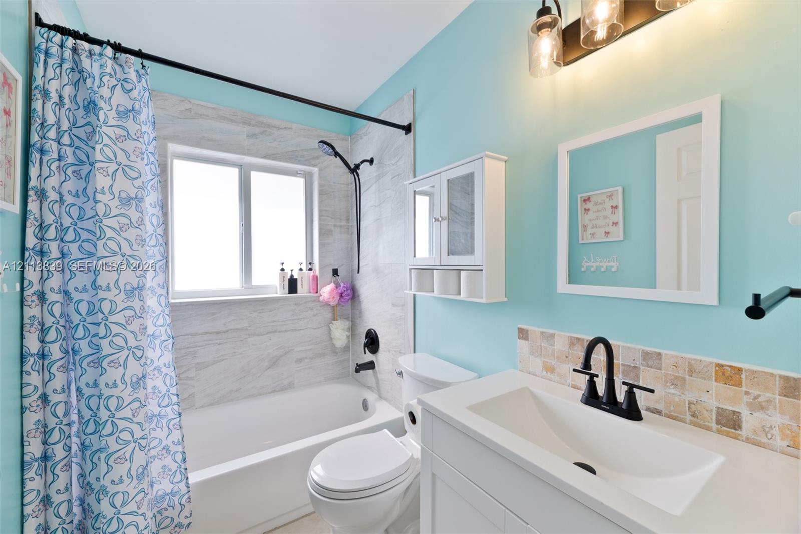 Bathroom featuring a vanity with an integrated sink, matte black hardware, stone-finish tile backsplash, bathtub with stone-finish tile surround, and wall-mounted cabinetry