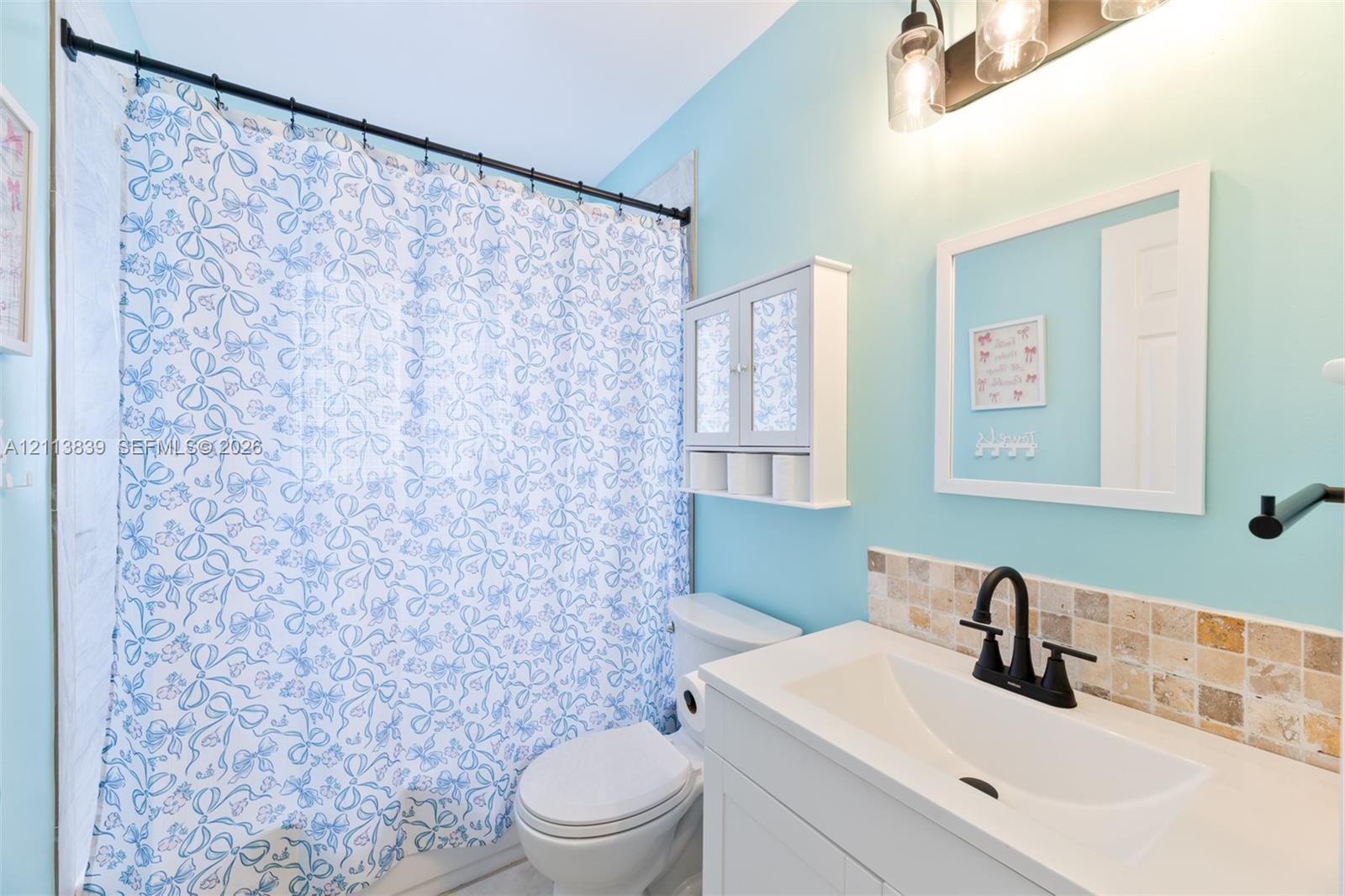 Bathroom featuring a vanity with a white countertop, matte black faucet, wall-mounted storage cabinet, and stone tile backsplash