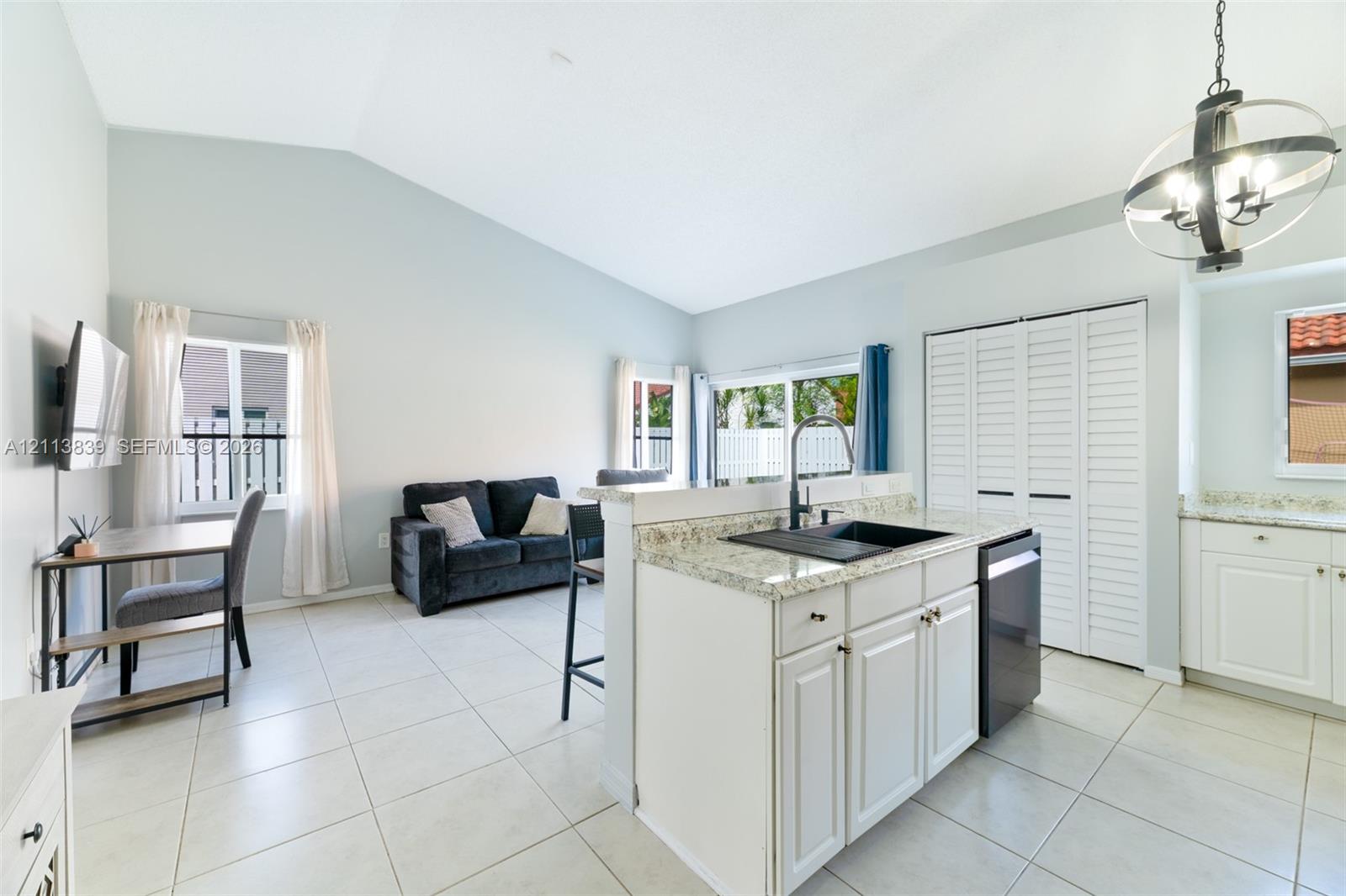 Vaulted ceilings, kitchen island with stone-surface countertop, dark composite undermount sink, white cabinetry with shaker-style doors, and large-format tile flooring