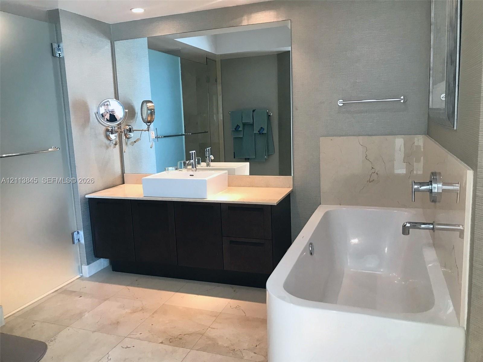 Dark wood-finish vanity with a rectangular vessel sink and countertop, large wall-mounted mirror, soaking tub with polished chrome fixtures, and large-format stone-finish flooring Dark wood-finish vanity with a rectangular vessel sink and countertop, large wall-mounted mirror, soaking tub with polished chrome fixtures, and large-format stone-finish flooring