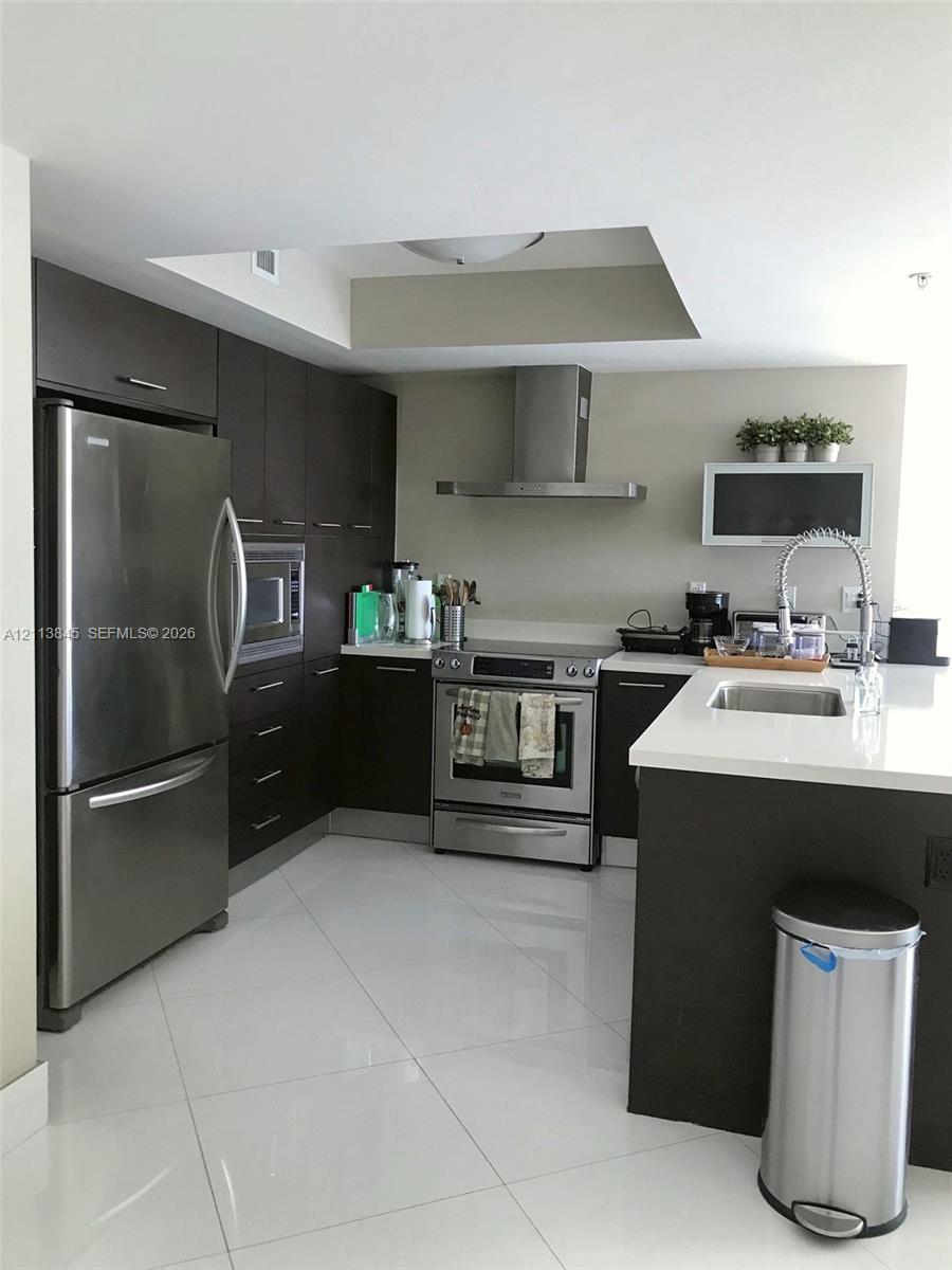 Kitchen featuring dark wood cabinetry, white stone-surface countertops, stainless steel appliances, a stainless steel chimney-style range hood, and polished tile flooring