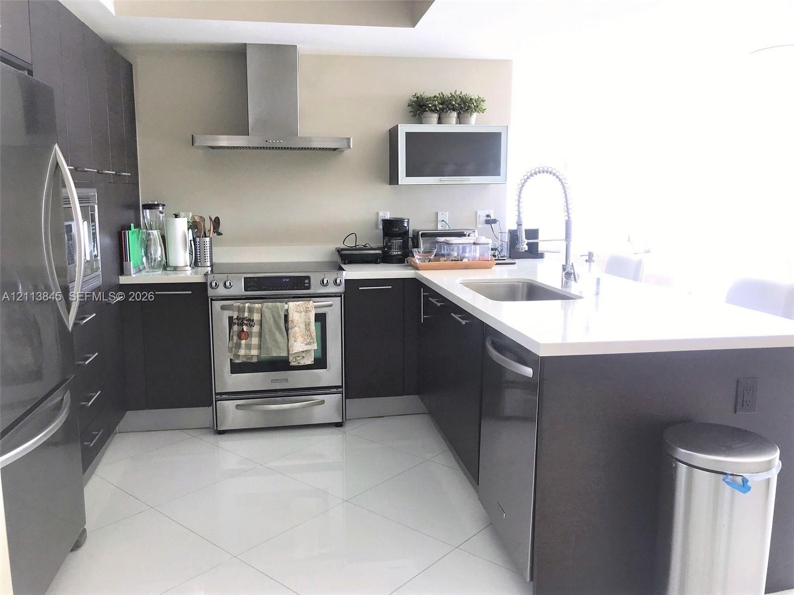 Kitchen featuring dark-finish cabinetry, stainless steel appliances, a chimney-style range hood, solid-surface countertops, and large-format tile flooring