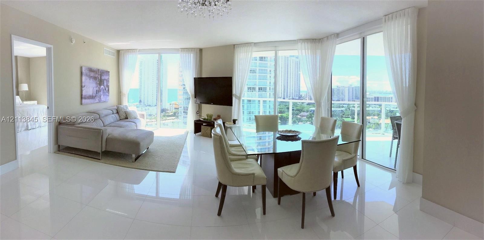 Open-concept living and dining area featuring polished tile flooring, floor-to-ceiling glass sliding doors, and a crystal chandelier Open-concept living and dining area featuring polished tile flooring, floor-to-ceiling glass sliding doors, and a crystal chandelier