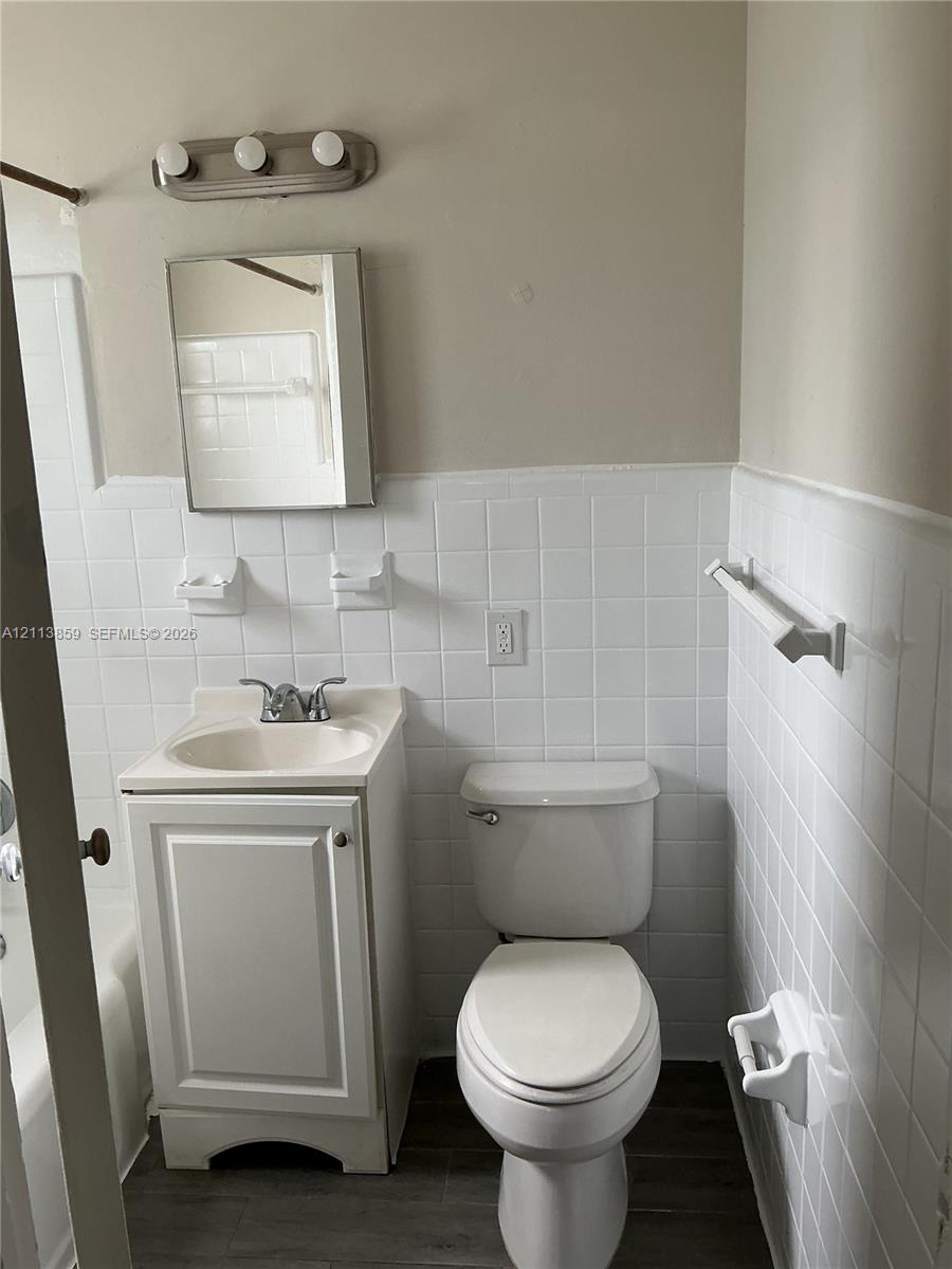 Bathroom featuring white ceramic wall tiling, a vanity with a drop-in basin and dual-handle faucet, a rectangular medicine cabinet, and wood-finish flooring