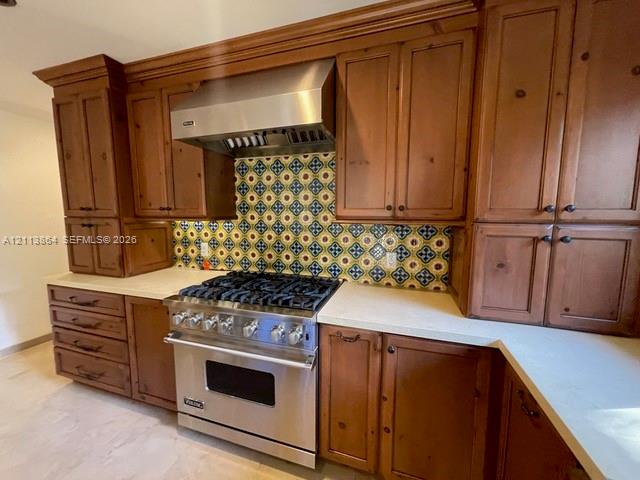 Kitchen featuring custom wood cabinetry, stainless steel professional-grade range, stainless steel vent hood, patterned tile backsplash, and light-toned countertops