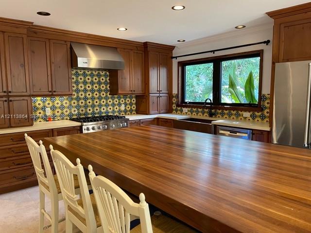 Kitchen featuring custom wood cabinetry, a large wood-topped center island, patterned tile backsplash, stainless steel range with a matching vent hood, and a deep apron-front sink