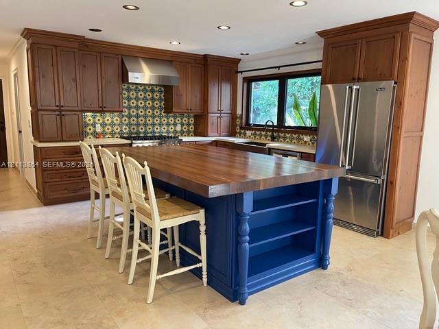 Kitchen featuring natural wood cabinetry, a central island with a wood countertop and open shelving, patterned tile backsplash, stainless steel appliances, and tile flooring