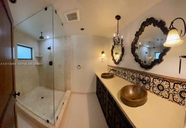 Double vanity featuring dual hammered metal vessel sinks, patterned tile backsplash, glass-enclosed walk-in shower with stone-finish surround, and decorative wall-mounted light fixtures