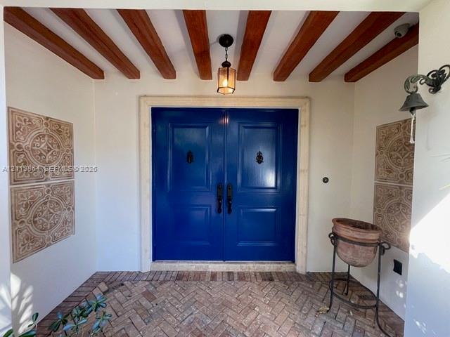Entryway featuring double solid-panel doors with iron knockers, exposed wood ceiling beams, decorative wall tile accents, and herringbone-patterned brick flooring