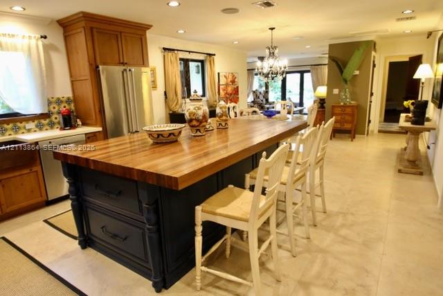 Kitchen featuring a large central island with a wood countertop, dark-finished cabinetry, stainless steel refrigerator, patterned tile backsplash, and light-toned tile flooring