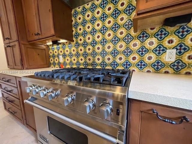 Stainless steel gas range with a heavy-duty burner grate, decorative multi-colored tile backsplash, solid-surface countertops, and wood-finish cabinetry featuring dark metal hardware