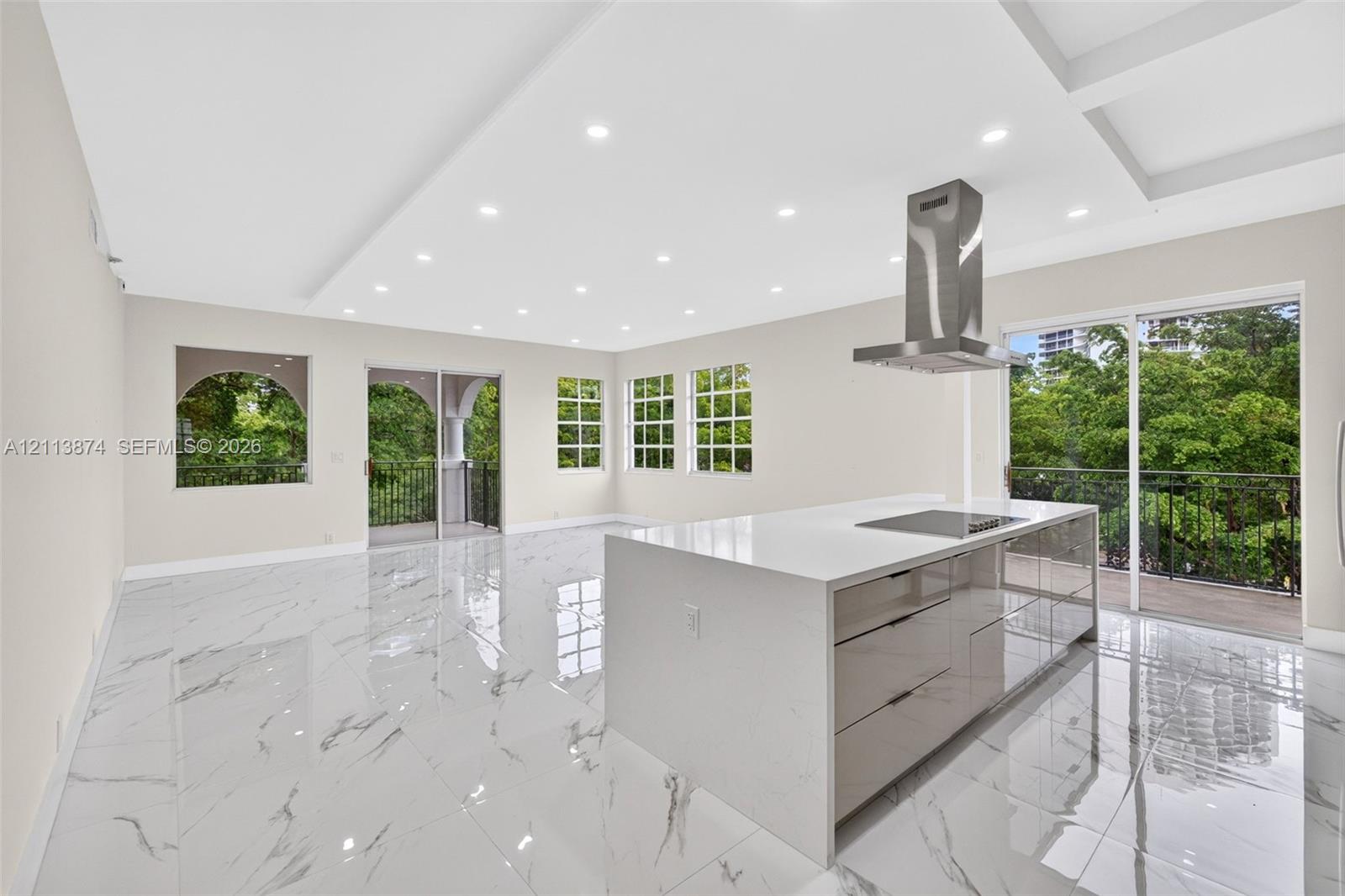Large-format stone-style flooring, central kitchen island with integrated cooktop, stainless steel range hood, recessed ceiling lighting, and multiple sliding glass doors