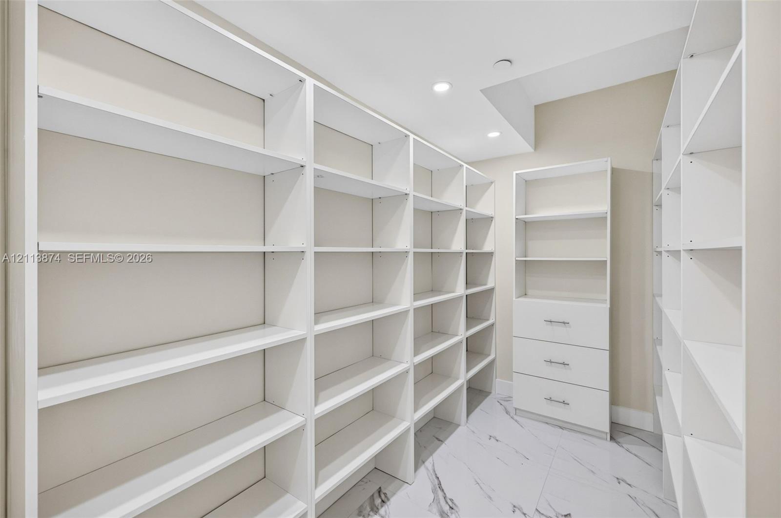 Custom walk-in storage featuring extensive floor-to-ceiling white shelving, a three-drawer built-in cabinet with metallic hardware, and large-format porcelain tile flooring