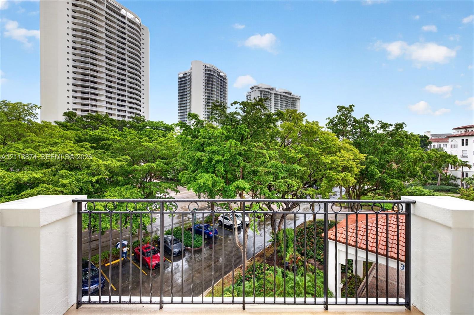 Balcony featuring a black metal railing and concrete pillars, overlooking mature trees and surrounding high-rise buildings