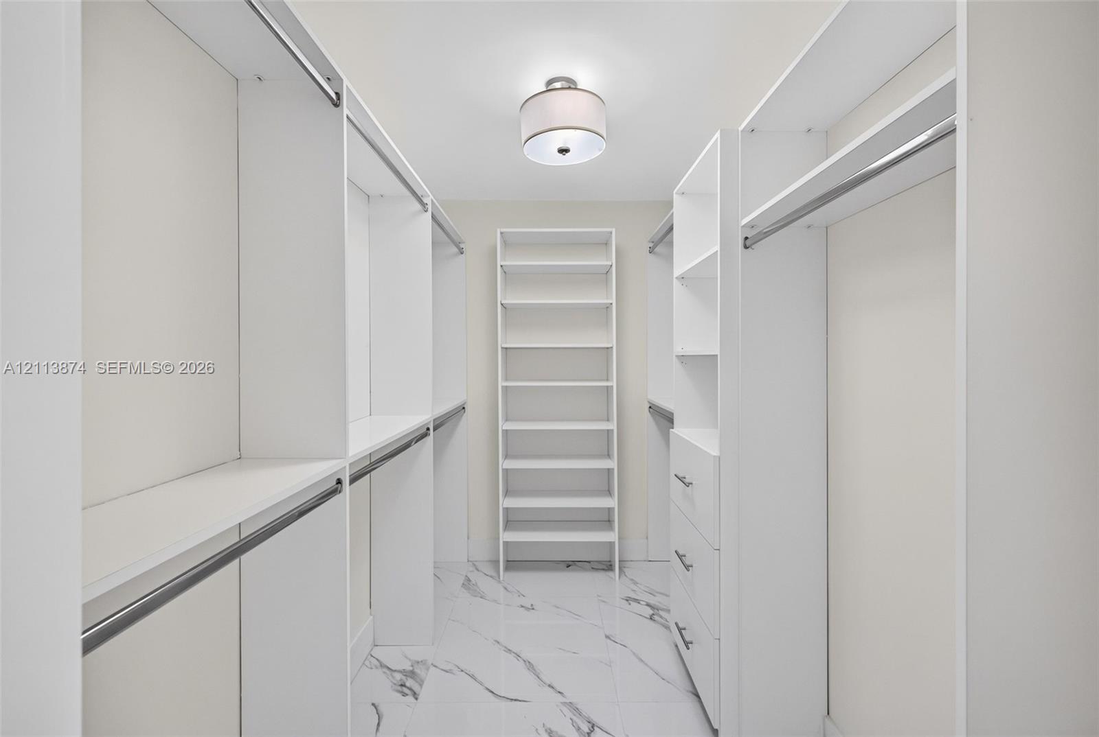Walk-in closet featuring white built-in shelving, integrated drawers, metal hanging rods, patterned tile flooring, and a drum-style ceiling light fixture