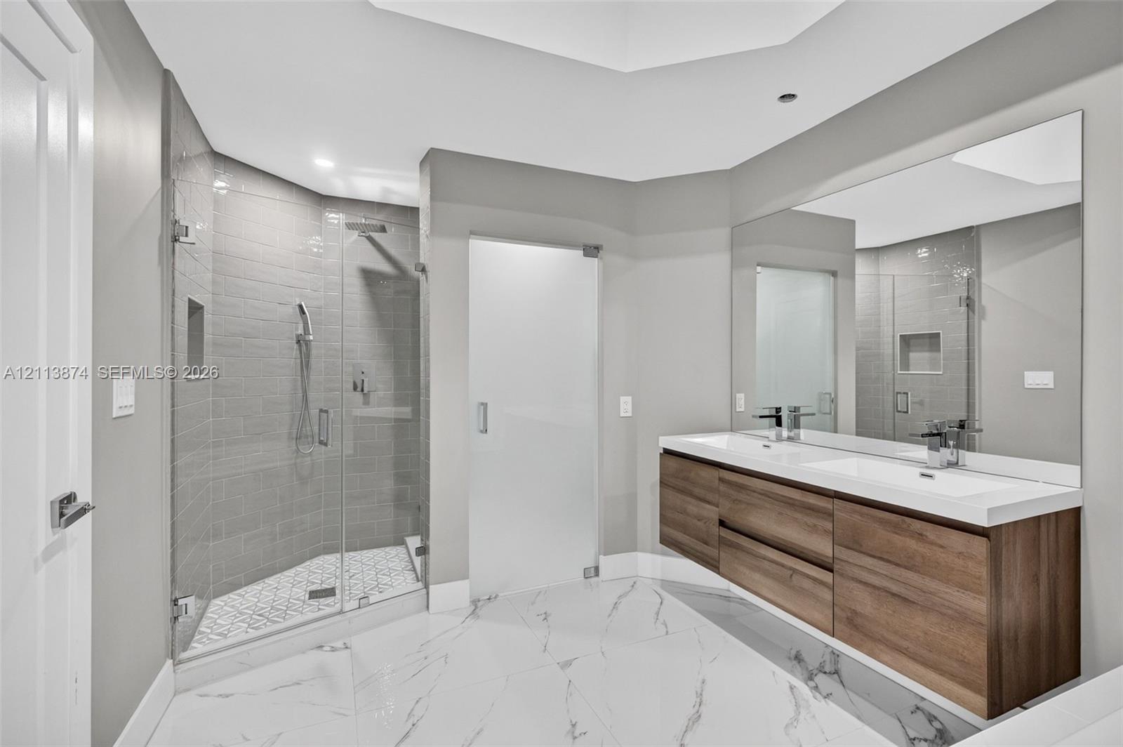 Floating dual-sink vanity with wood-finish cabinetry, frameless glass shower enclosure featuring tiled walls, and large-format floor tiling